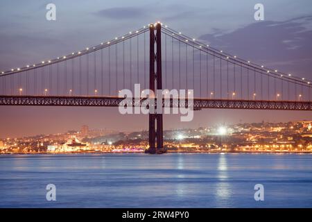 25 avril (portugais : Ponte 25 de Abril) Pont sur le Tage la nuit et ville de Lisbonne, Portugal en arrière-plan Banque D'Images