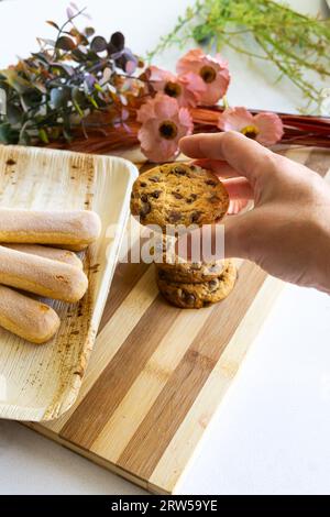 Main de femme tenant un biscuit maison avec des pépites de chocolat d'une table décorée. Tir vertical. Banque D'Images