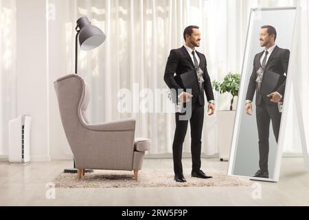 Homme d'affaires debout avec une mallette pleine d'argent à la maison et regardant dans un miroir Banque D'Images