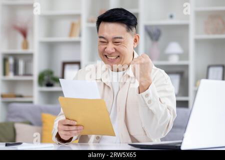 Heureux et réussi asian a reçu l'enveloppe de notification de courrier avec de bonnes nouvelles, homme assis à table à la maison, travaillant dans le bureau à domicile dans le salon, célébrant joyeusement les nouvelles gagnantes. Banque D'Images