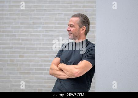 Homme attrayant aux cheveux gris se penche contre un mur avec ses bras croisés et regarde pensivement dans la distance en face d'un mur de briques blanches Banque D'Images