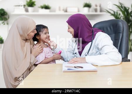 Souriant jeune médecin musulman portant le hijab vérifiant la fille à la clinique moderne. Pédiatre féminin amical examiner le patient jeune enfant avec sa mère Banque D'Images