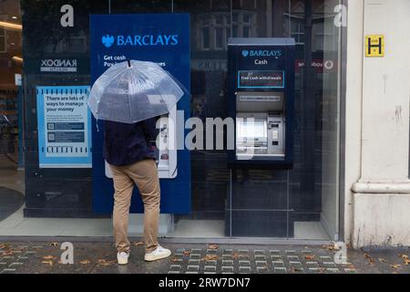 Homme tenant un parapluie retirant de l'argent d'un distributeur de billets de banque Barclays, High Street Kensington, Londres, Angleterre, Royaume-Uni Banque D'Images