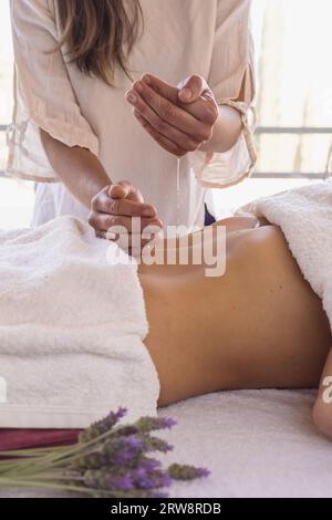 Gros plan de thérapeute féminin appliquant de l'huile de massage sur les mains avant la thérapie sur la dame allongée sur la table de massage. Banque D'Images