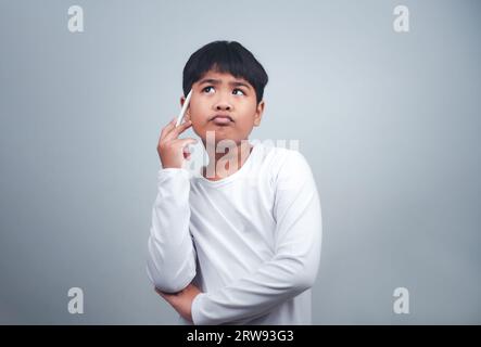 Un garçon en chemise blanche tient un stylo blanc sur fond blanc. Fait preuve de réflexion, de réflexion et d'examen des options. Banque D'Images