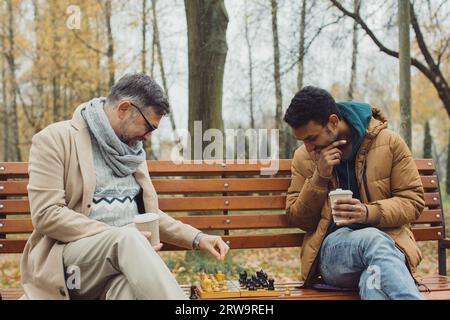 Des amis jouent aux échecs sur un banc dans un parc d'automne. Amitié multiculturelle de personnes d'âges différents Banque D'Images