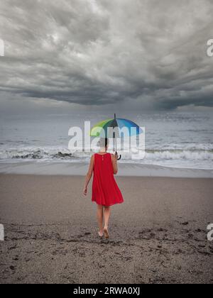 Une fille dans une robe rouge sur le bord de la mer avec un parapluie multicolore Banque D'Images