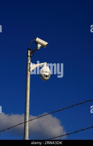 Deux caméras de surveillance sur un poteau derrière des barbelés, fond de ciel bleu avec des nuages Banque D'Images