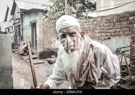 Khulna, Bangladesh, vers juillet 2012 : un vieil homme en vêtements blancs et bonnet blanc avec une longue barbe blanche tient un bâton de bois dans un bidonville à Khula, Bangladesh. Banque D'Images