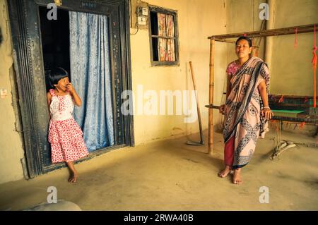 Srimongal, Bangladesh, vers juillet 2012 : une femme autochtone en écharpe se penche sur un métier à tisser avec sa fille souriante en robe rouge et blanche à Srimongal Banque D'Images