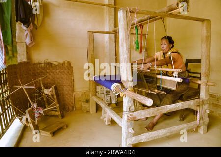 Srimongal, Bangladesh, vers juillet 2012 : une femme aux cheveux noirs pieds nus avec une écharpe jaune est assise sur une chaise en bois et tisse avec un tissu bleu à Srimongal Banque D'Images