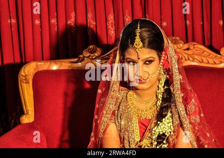 Dhaka, Bangladesh, vers juillet 2012 : jeune mariée aux cheveux noirs dans une belle robe scintillante rouge et or est assise sur un canapé rouge et regarde droit à Dhaka Banque D'Images