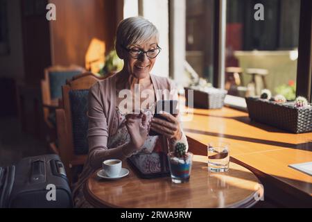 Femme mature est assise dans un café à l'aéroport, attendant l'embarquement. Elle examine l'horaire sur smartphone. Banque D'Images