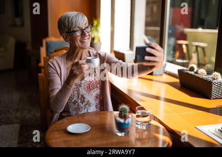 Femme mûre boit du café dans un café. Elle prend un selfie. Banque D'Images