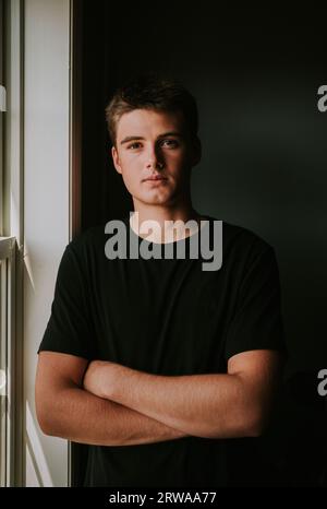 Portrait de beau garçon adolescent debout à côté de la fenêtre dans la chambre sombre. Banque D'Images
