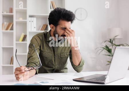 L'homme indien fatigué enlève des lunettes, souffre de sensations douloureuses, a des problèmes d'astigmatisme. Un homme stressé après une longue journée de travail au Banque D'Images