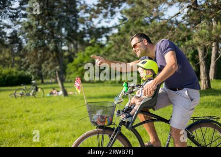 Regarde là-bas. Journée familiale active dans la nature. Père et fils font du vélo à travers le parc de la ville le jour d'été unny. Un garçon mignon est assis dans la chaise de vélo avant tandis que le père fait du vélo. Père fils de liaison Banque D'Images