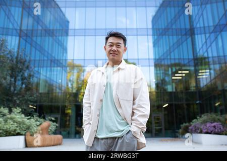 Portrait d'un jeune homme d'affaires asiatique, programmeur, freelance debout dans un centre de bureau extérieur, tenant ses mains dans ses poches et regardant avec confiance la caméra. Banque D'Images