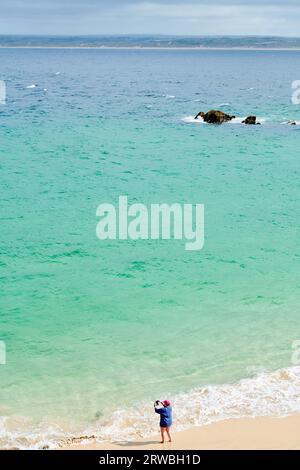 Un touriste solitaire prend une photo sur la plage de sable de Bamaluz à St Ives, Cornouailles, tandis que la marée de l'océan Atlantique est dehors par une journée nuageuse. Banque D'Images