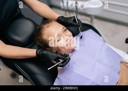 Vue à grand angle de mignon petite fille souriant regardant loin assis dans le siège de stomatologie tandis que dentiste pédiatrique dans des gants prêt à faire l'examen des dents. Banque D'Images