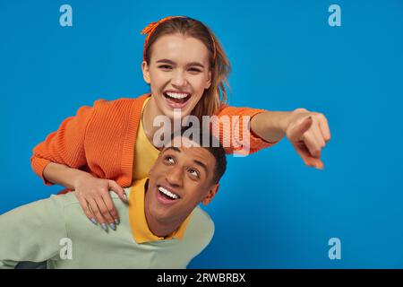 joyeux homme afro-américain piggybacking joyeuse petite amie sur fond bleu, pointant vers la caméra Banque D'Images