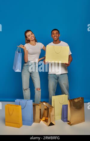 joyeux couple multiethnique dans une tenue décontractée tenant des sacs à provisions sur fond bleu, achat frénétique Banque D'Images