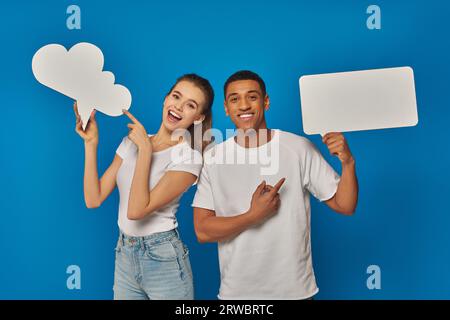 couple interracial étonné pointant des pancartes vierges sur fond bleu, la pensée et les bulles de parole Banque D'Images