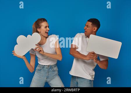 couple interracial émotionnel tenant des pancartes vierges sur fond bleu, des bulles de pensée et de parole Banque D'Images