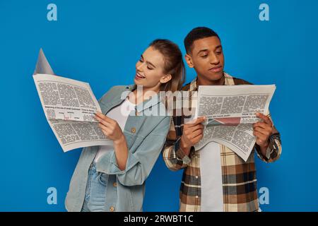 heureux homme afro-américain et jolie femme caucasienne lisant des nouvelles sur fond bleu, journaux Banque D'Images