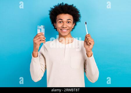 Portrait de la belle personne joyeuse les bras de sourire toothy tiennent l'eau en verre de brosse à dents isolée sur fond de couleur bleue Banque D'Images