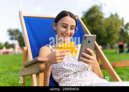Femme ravie dans des lunettes assis dans une chaise avec carte de crédit en main dans le parc et shopping en ligne avec téléphone portable Banque D'Images