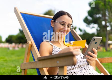 Femme ravie dans des lunettes assis dans une chaise avec carte de crédit en main dans le parc et shopping en ligne avec téléphone portable Banque D'Images
