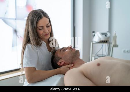 Positif jeune femme thérapeute en uniforme blanc souriant et massant cou d'un patient mâle calme allongé sur la table sans chemise pendant les ses de physiothérapie Banque D'Images