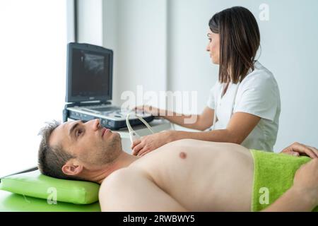 Calmer jeune thérapeute féminine en uniforme blanc en utilisant la machine à ultrasons professionnelle pour vérifier l'épaule du patient masculin pendant le rendez-vous dans clin moderne Banque D'Images