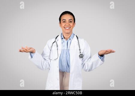 Souriante jeune femme médecin latine tenant quelque chose d'invisible sur ses mains, posant sur fond gris clair de studio Banque D'Images