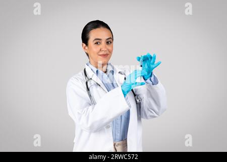 Jeune femme brésilienne médecin en manteau blanc et gants prenant le vaccin à la seringue, posant sur fond gris Banque D'Images