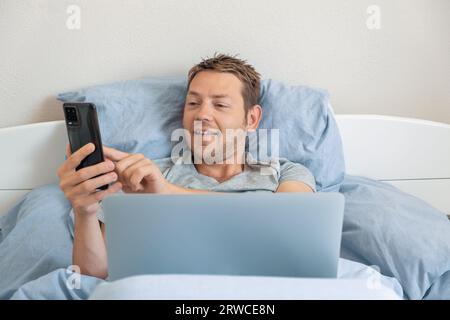 Homme caucasien joyeux souriant et envoyant un message texte sur smartphone pendant le chat vidéo sur ordinateur portable. Homme couché dans le lit et ayant la conversation en ligne dans Banque D'Images