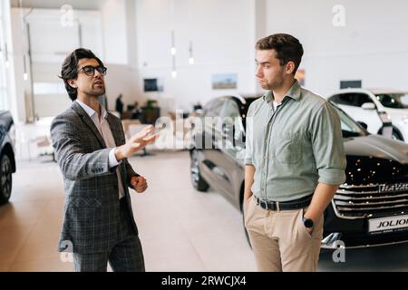 Photo moyenne d'un concessionnaire automobile masculin compétent en costume d'affaires ayant une conversation avec un client jeune homme sérieux à la salle d'exposition. Client douteux Banque D'Images