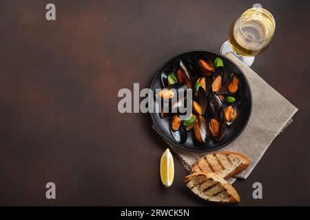 Moules de fruits de mer et feuilles de basilic dans une assiette noire avec verre de vin, tranches de pain, toile de jute sur fond rouillé. Vue de dessus avec emplacement pour votre texte. Banque D'Images