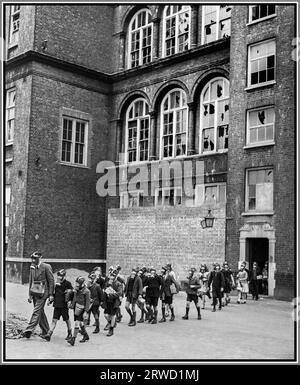 LA foreuse anti-gaz BLITZ LONDON WW2 à Old Woolwich Road School à Greenwich, Londres 1941. Un enseignant portant un masque à gaz conduit un groupe d'écoliers hors de l'entrée de l'école Old Woolwich Road School à Greenwich, Londres. Les enfants traversent le terrain de jeux, tous portent leurs masques à gaz. L'école a clairement été endommagée par les raids aériens blitz de l'Allemagne nazie, de nombreuses fenêtres ont des vitres manquantes ou brisées. 1941 Londres Blitz bataille d'Angleterre Seconde Guerre mondiale Banque D'Images