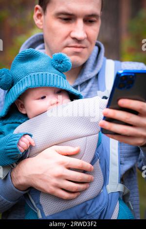 Père et bébé nouveau-né dans une fronde dans la forêt d'automne, homme avec smartphone. Famille heureuse. Banque D'Images