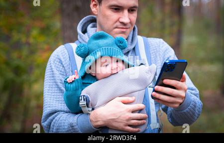 Père et bébé nouveau-né dans une écharpe dans la forêt d'automne, homme avec téléphone portable. Famille heureuse. Banque D'Images