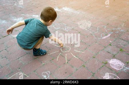 Petit garçon de deux ou trois ans dessine le soleil avec des crayons dans le parc sur des tuiles. Temps de jeu pour les enfants. Jeux pour enfants. Mise au point sélective Banque D'Images