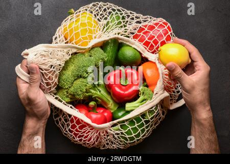 Homme mettant le citron dans un sac en maille avec différents légumes frais sur fond noir Banque D'Images