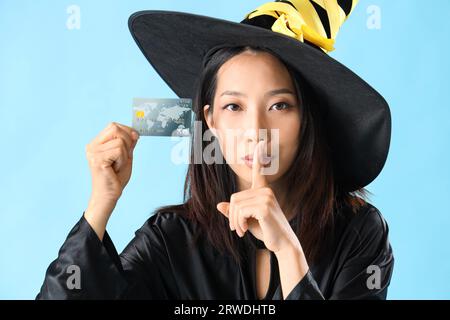 Jeune femme asiatique habillée pour Halloween comme sorcière avec carte de crédit montrant geste de silence sur fond bleu, closeup Banque D'Images