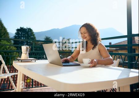 Souriant heureuse entrepreneure, agent immobilier sourit en regardant le moniteur d'ordinateur portable, profitant de sa pause café tout en travaillant en ligne dans le terr à la maison Banque D'Images