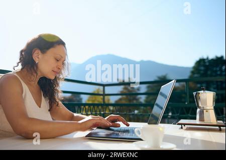 Confiante femme d'affaires d'âge moyen assise à table dans la terrasse de la maison, travaillant à distance sur ordinateur portable, tapant du texte, planifiant un nouveau projet d'entreprise, livre Banque D'Images