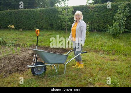 femme fermière senior des années 50 avec une pelle décharge une brouette remplie de terre. Travaux sur le terrain à ressort Banque D'Images