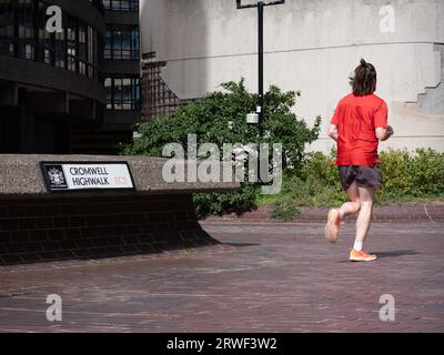 Coureur passant devant le panneau Cromwell Highwalk, The Barbican Estate, ou Barbican, quartier résidentiel d'environ 2 000 appartements, maisonnettes et maisons dans la City of London, Angleterre, avec mur en ruine en arrière-plan Banque D'Images