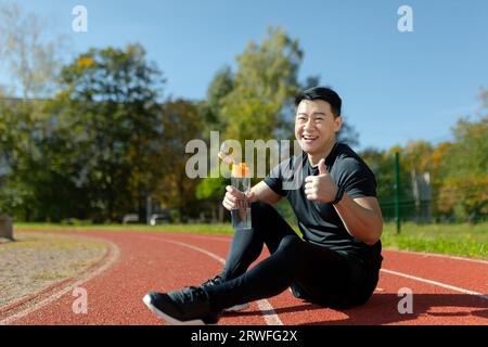 Fatigué et souriant jeune coureur masculin asiatique assis et reposant sur la piste du stade, tenant la bouteille d'eau et montrant un super geste avec le doigt. Banque D'Images